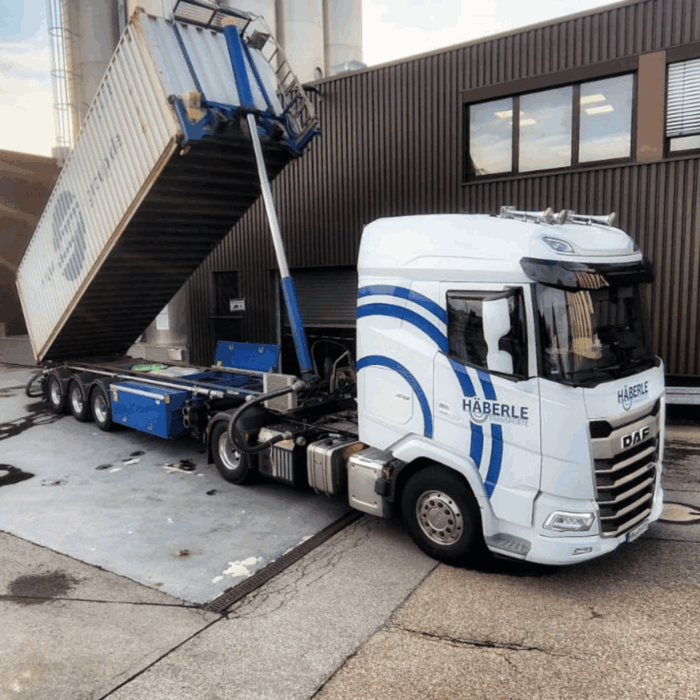 Weißer Lastwagen mit blauen Streifen kippt einen Container auf einem Industriegelände ab.