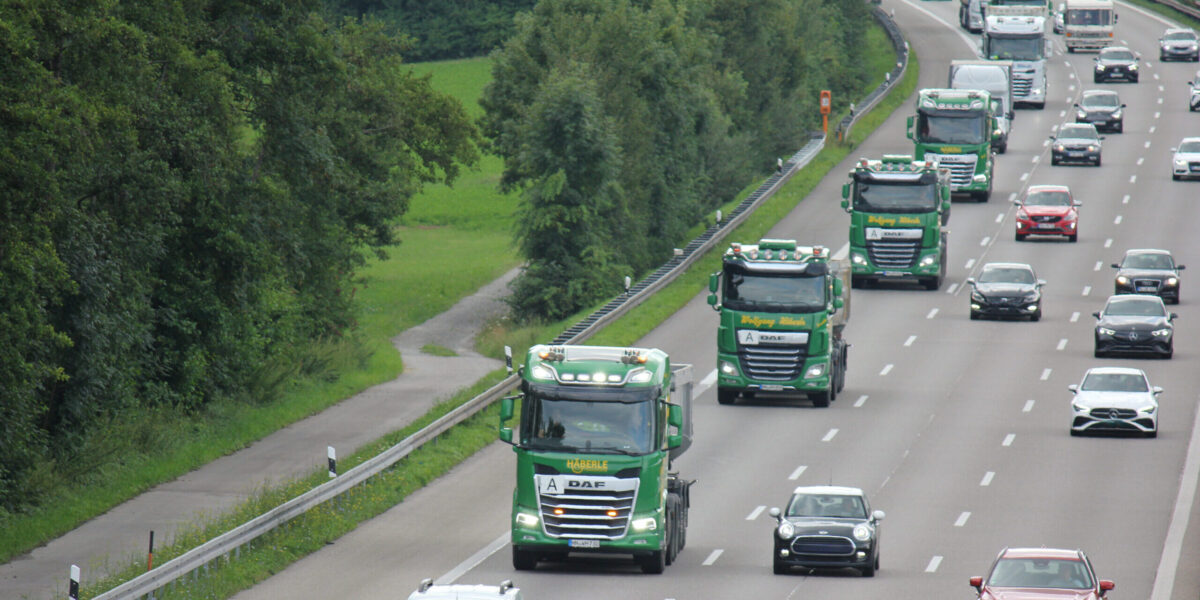 Mehrere grüne Lastwagen und Autos fahren auf einer stark befahrenen Autobahn, umgeben von grünen Bäumen.